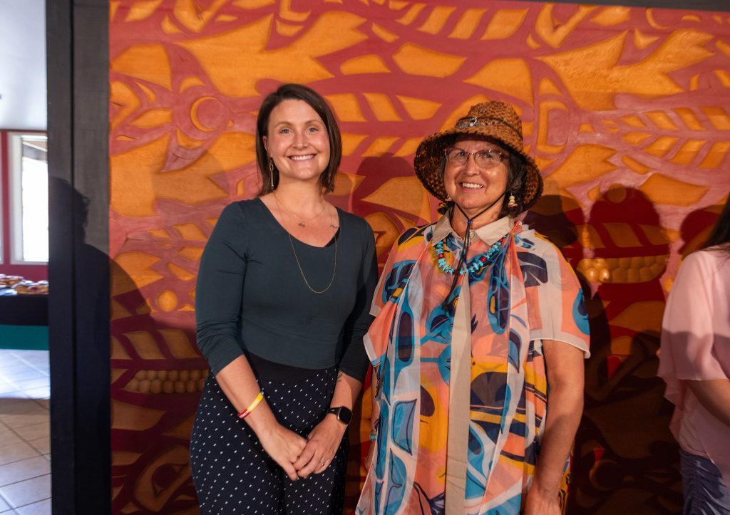 A woman in a green top smiles alongside a woman in Quw'utsun regalia in front of background of Indigenous art.