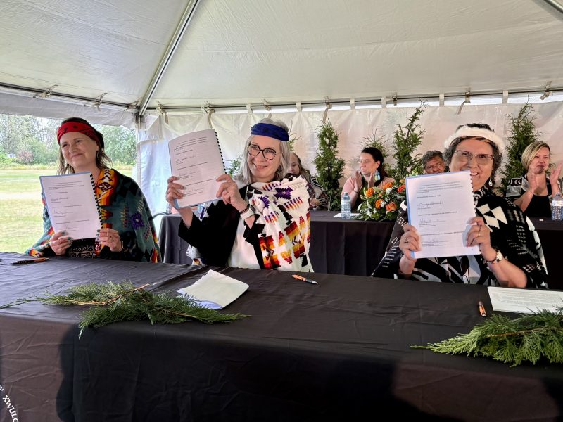 B.C. Minister of Children and Family Development, Grace Lore; Federal Minister of Indigenous Services, Patty Hajdu; and Quw’utsun Chief Cindy Daniels hold up the signed agreements from a table on a stage. They smile for the cameras.