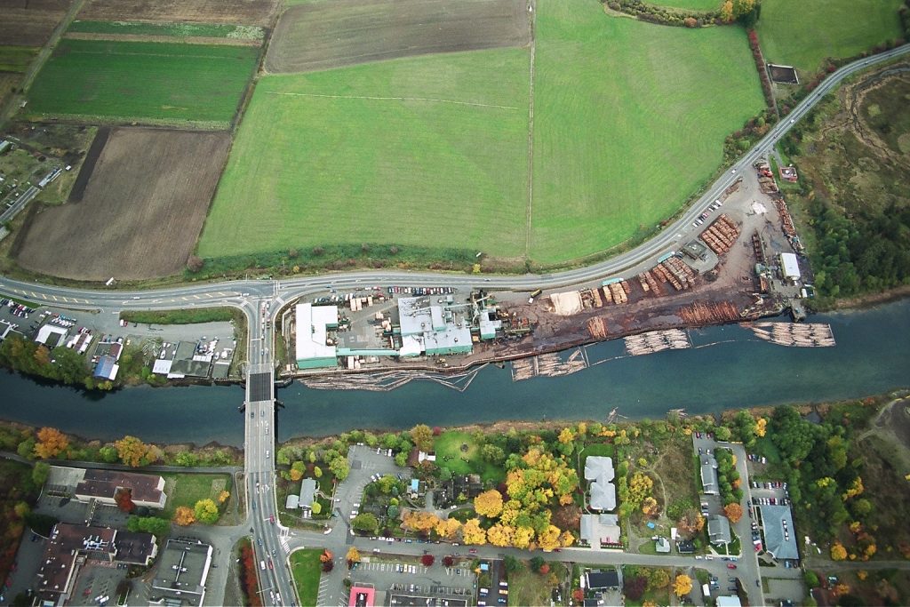 An aerial view of the Field Sawmill alongside the estuary. A log boom is seen alongside the side of the water, and cars are crossing a bridge next to the site. A farm field sits in the background, and trees and houses in the foreground.