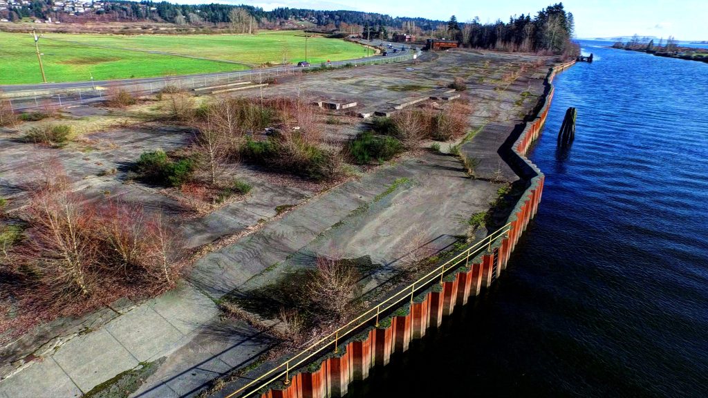 Aerial view of an estuary and an abandoned industrial site on the bank. A steel wall is seen alongside the shoreline. The site is mostly concreate with some bushes growing though.