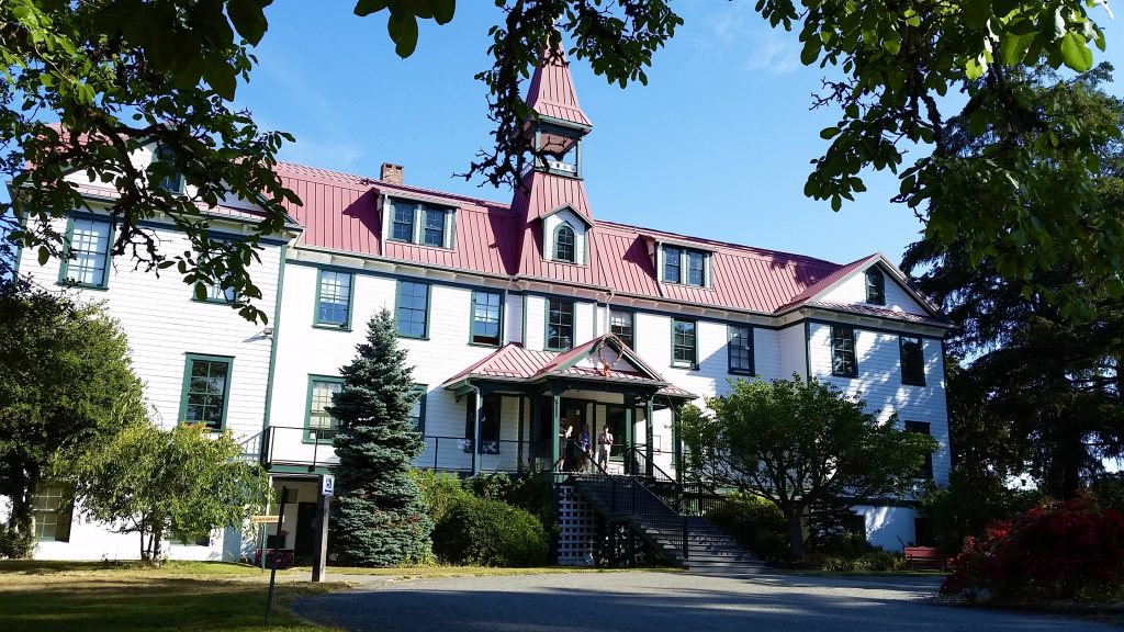 A white, two-storey old school building with a red roof stands under a blue sky.
