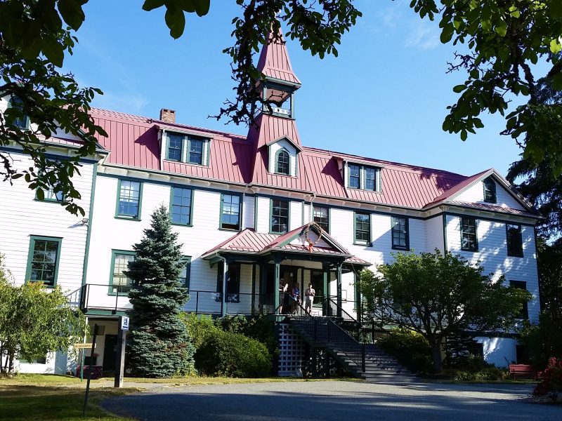A white, two-storey old school building with a red roof stands under a blue sky.