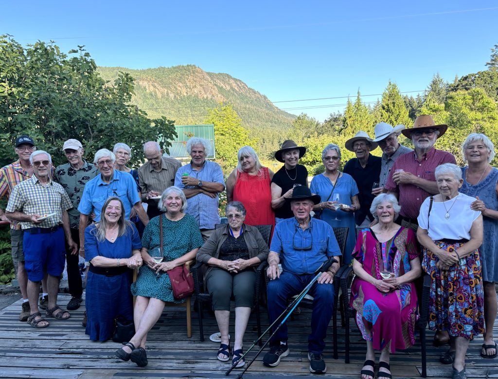 Eighteen people sit and stand in a group, posing for a picture in front of a scenic valley and mountain. They are on Providence Farm.
