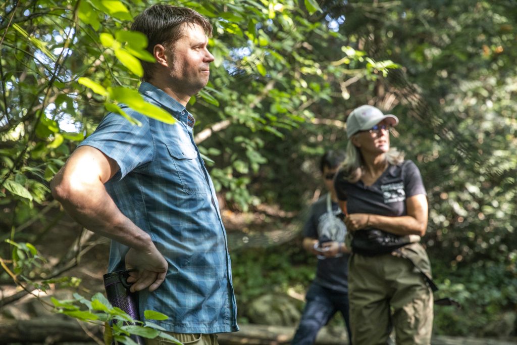 A man in the foreground stands at a creek lined with green trees. A woman in a baseball cap and man stand further up the river as they study streams for a chemical known as 6PPD-q