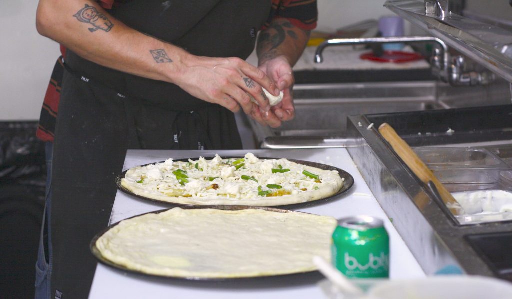 A pair of tattooed hands sprinkle cheese on a pizza dough