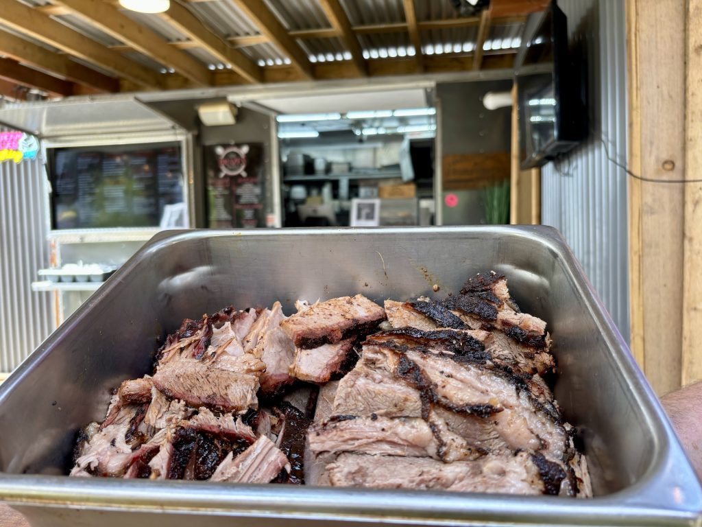 A metal pan full of smoked brisket chunks is displayed in front of a kitchen window of a food truck.