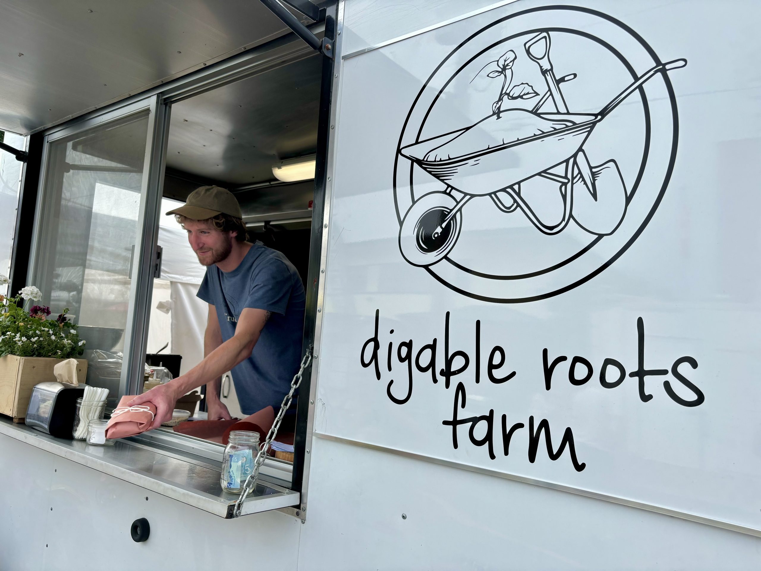 A man in a baseball cap hands a sandwich wrapped in butcher paper to an unseen customer from a white food truck window.