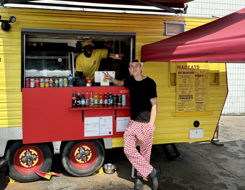 A young man in a tee shirt and checkered chef pants stands outside a yellow food trailer. Another employee smiles from the truck window.