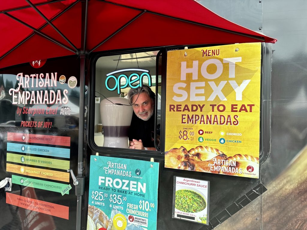 An older man with a grey beard looks out of the window of his food truck that is decorated in colourful signs promoting his empanadas.