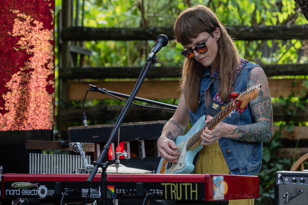MJ McGregor plays a guitar while standing in front of a keyboard with the word TRUTH written on it in a backyard. 