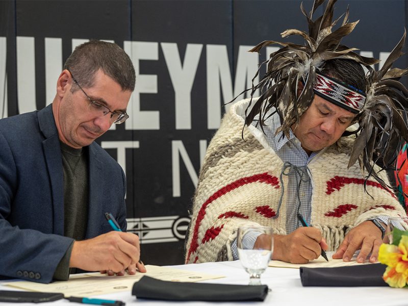 Photo of two people sitting beside each other, one wearing a blue blazer and the other traditional regalia and a button down shirt, signing an agreement.