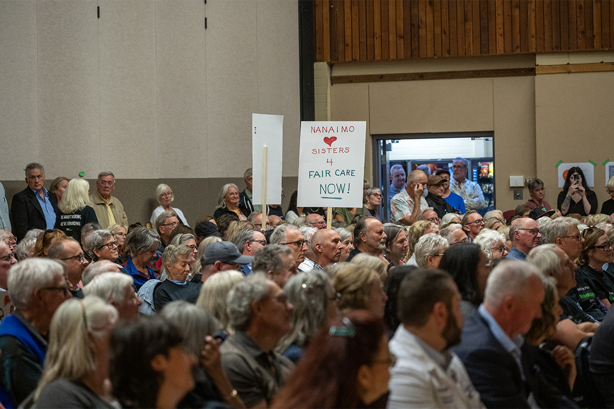 A large crowd is seated with many people standing around the crowd as well. A sign sticks up from the crowd that reads "Nanaimo sisters 4 fair care now!"