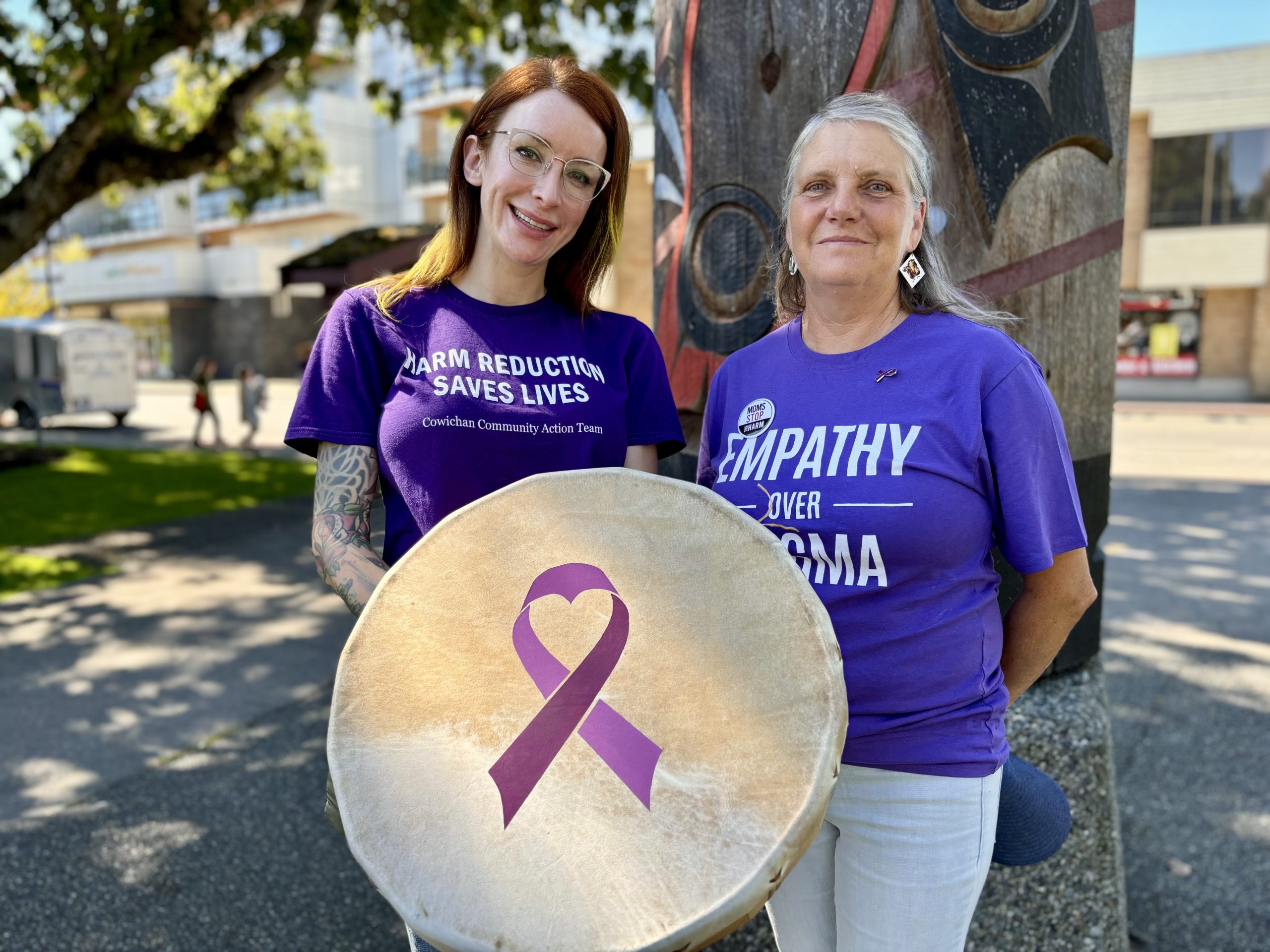 Two women wearing purple shirts for international overdose awareness day hold a drum that has a purple ribbon on it.