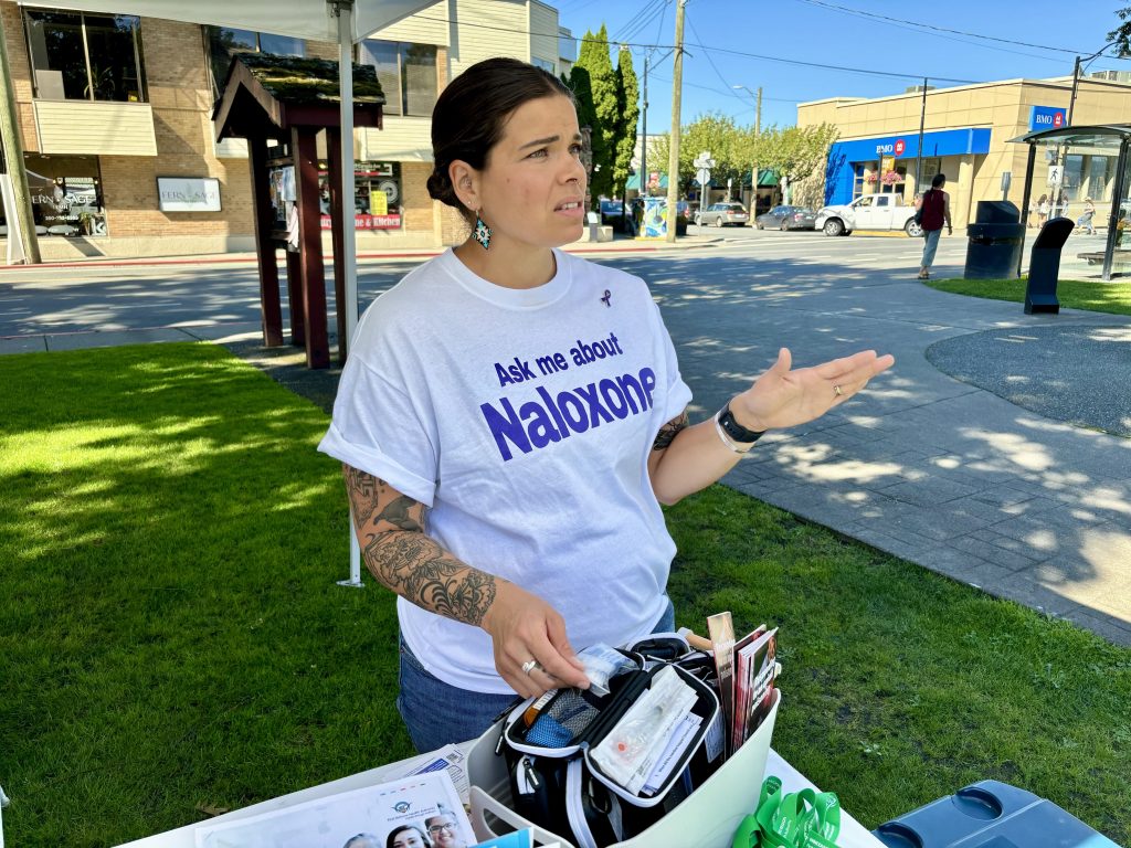 A woman wears a shirt that says "ask me about naloxone" and is speaking, with a naloxone kit in hand.