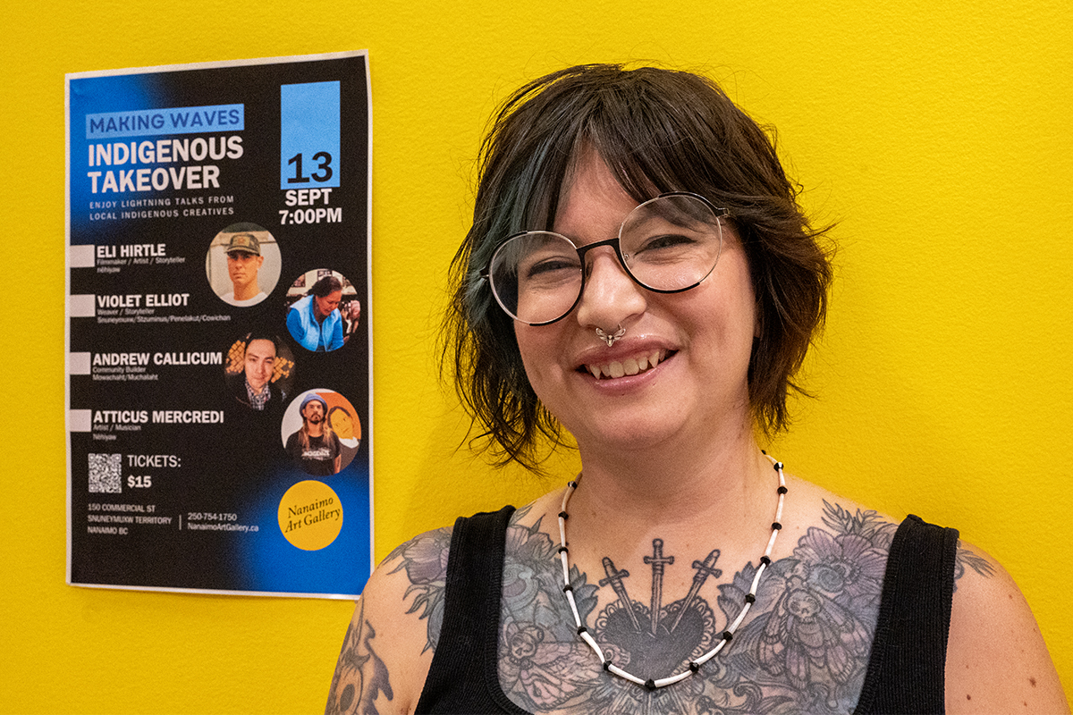 Photo of a person with short brown and coloured hair wearing glasses and smiling with visible tattoos on their chest. They are standing in front of a yellow wall with a poster for a Making Waves Indigenous Takeover event beside them.