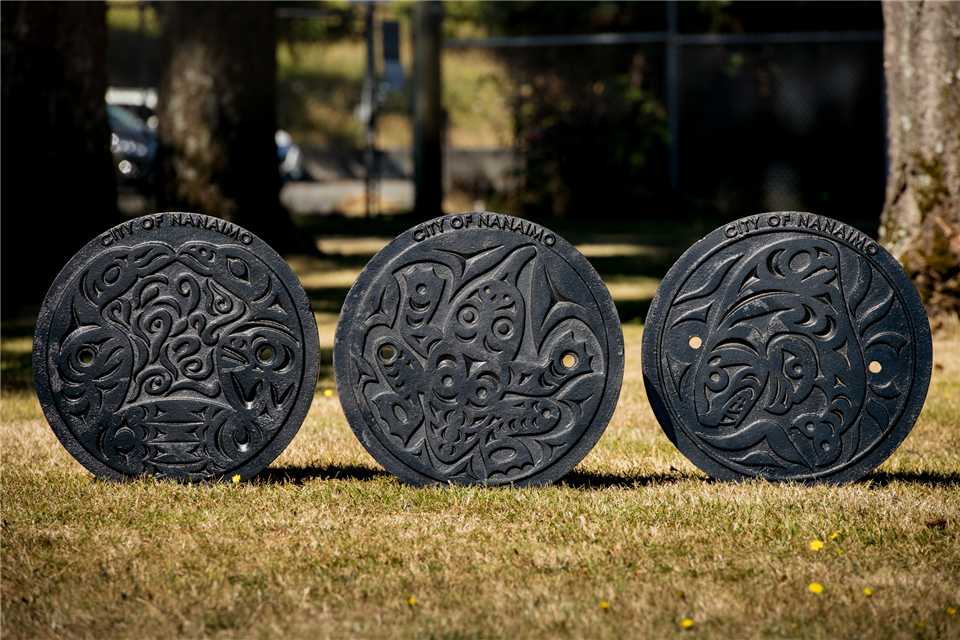 Three manhole covers stand up in grass side-by-side with designs of eagles, a frog and an orca by Joel Good.