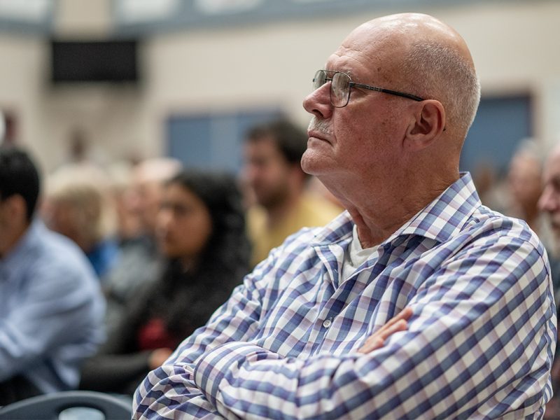A man wearing a blue and white checkered shirt sits in an audience with his arms crossed listening.
