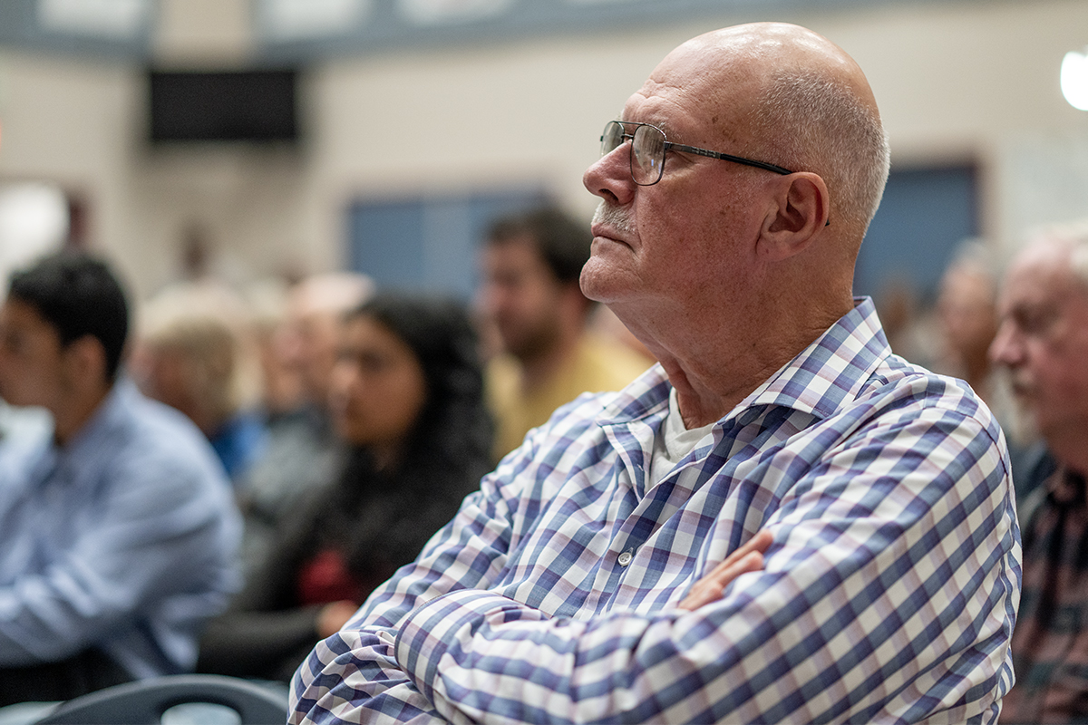 A man wearing a blue and white checkered shirt sits in an audience with his arms crossed listening.
