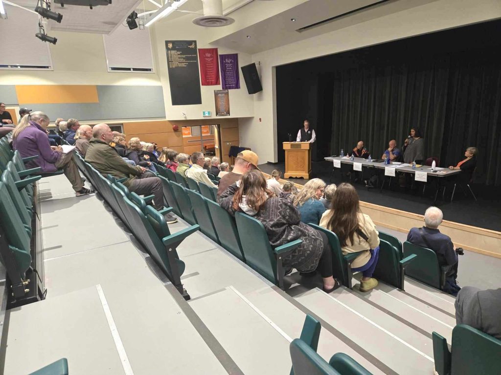 Bleachers with a few dozen people look down on a table of four candidates sitting at a table, with an MC standing behind a podium.