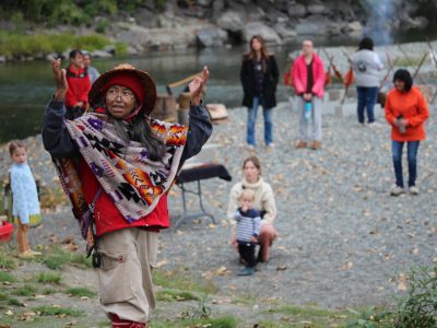 Della Rice Sylvester calls people together at Quw'utsun Sta'lo' Skweyul (Cowichan River Day). She is wearing a cedar hat and has arms in the air as a crowd gathers behind, on the riverbank.