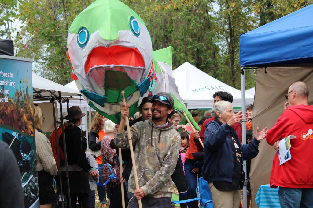 A giant salmon puppet is held by a line of people who are holding it up with sticks and walking and smiling.